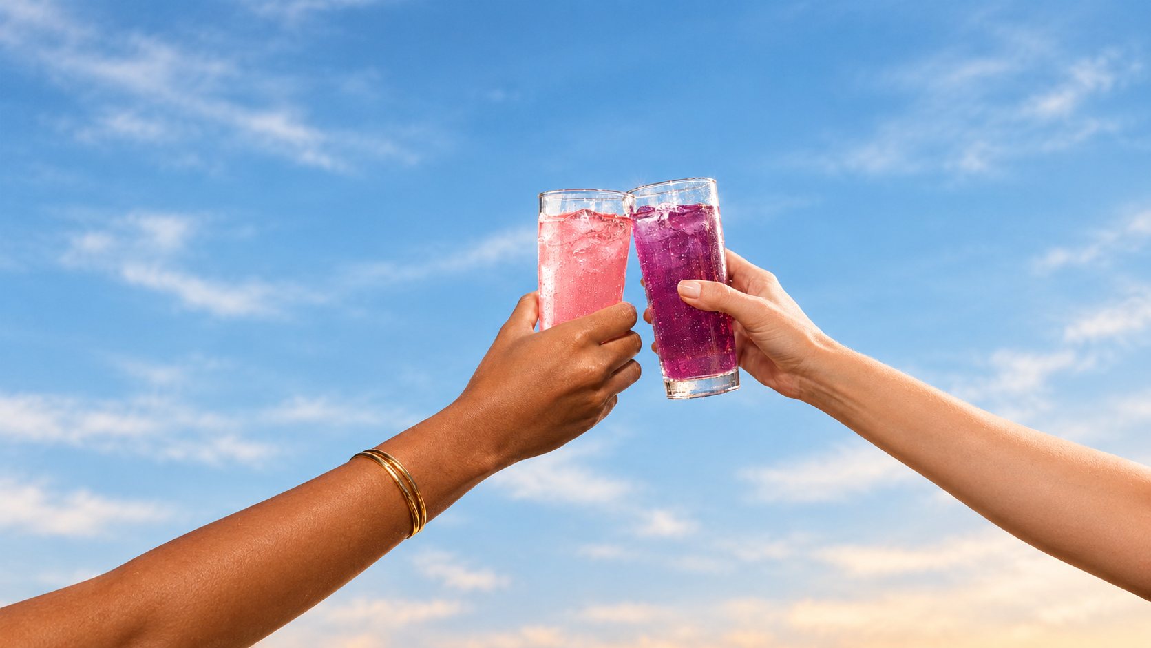 Dua tangan bersulang dengan minuman KukuBima merah muda dan ungu di bawah langit biru