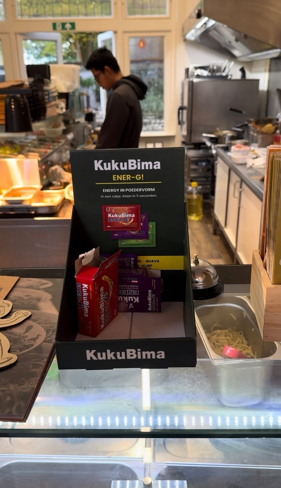 Display KukuBima berisi kotak dan sachet di meja kasir Jajanan, dengan dapur di latar belakang